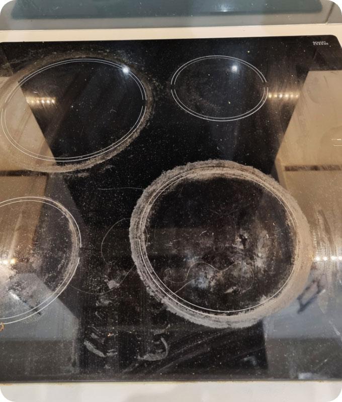 An image of a dirty black ceramic stove top covered in burnt food residue and greasy stains. The surface is marred with dried-on spills, splatters, and grime, making it appear heavily soiled and neglected. The burners and knobs are coated with grease and food remnants, and the entire stove top has a dull and unclean appearance.