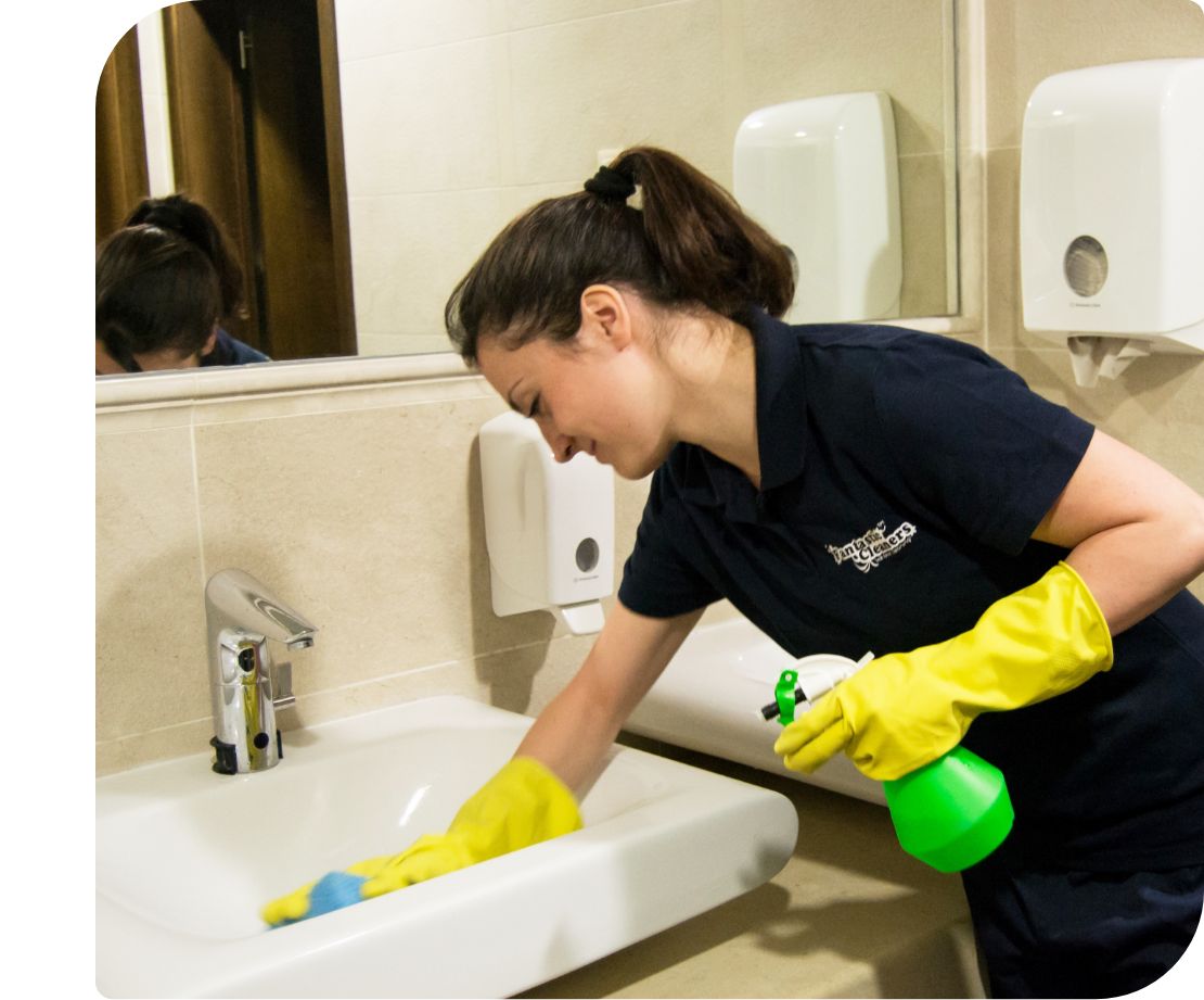 A professional cleaner in a dark uniform and yellow gloves cleans a sink using a spray bottle and a cloth.